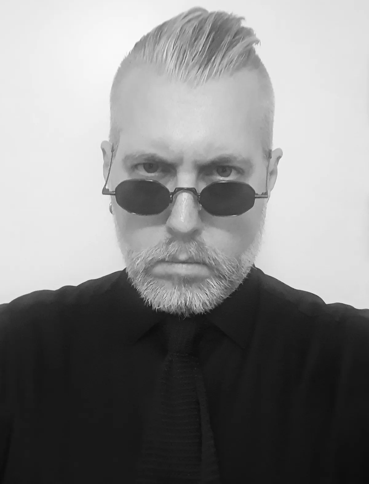 A black and white selfie of a man with a shaved sides haircut, a full beard, and small round sunglasses, wearing a black…