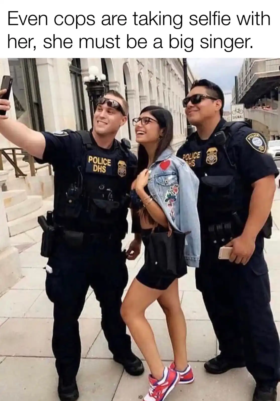 Three people, two police officers and a woman, are posing for a selfie on a city sidewalk. The officer on the left holds a…