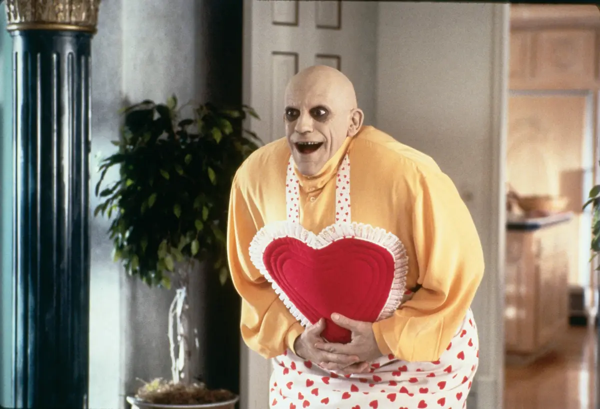 A bald man with exaggerated makeup and a wide, manic grin wears a yellow shirt and a white apron covered in red hearts. He…
