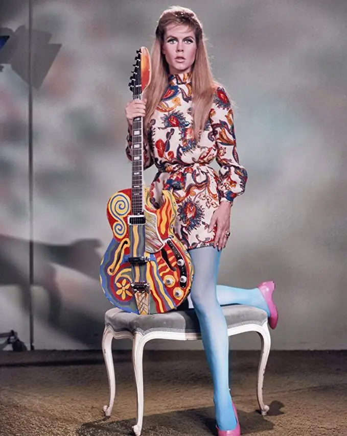 a woman in a psychedelic-patterned dress and blue tights sits on a chair holding a vibrantly painted electric guitar. Her…