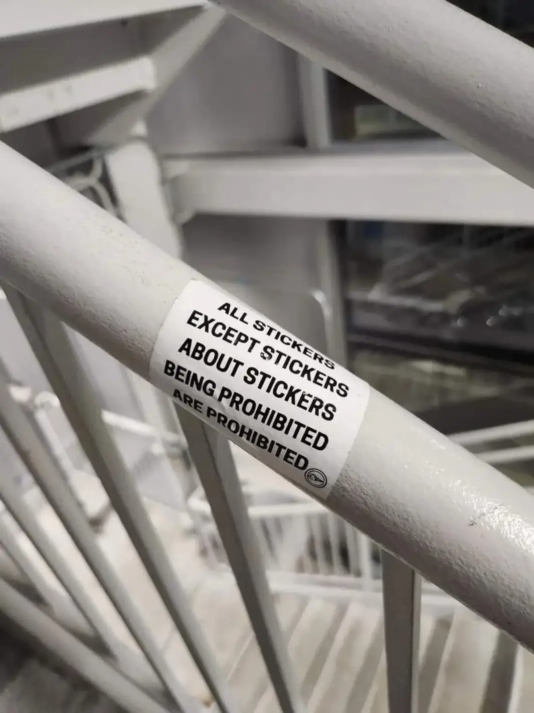 A close-up photo shows a white sticker on a white metal railing, which reads: 'ALL STICKERS EXCEPT STICKERS ABOUT STICKERS…
