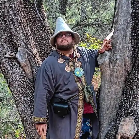 A man with a beard and glasses wears a tall, pointed wizard hat and a dark robe with ornate patterns and multiple necklaces…