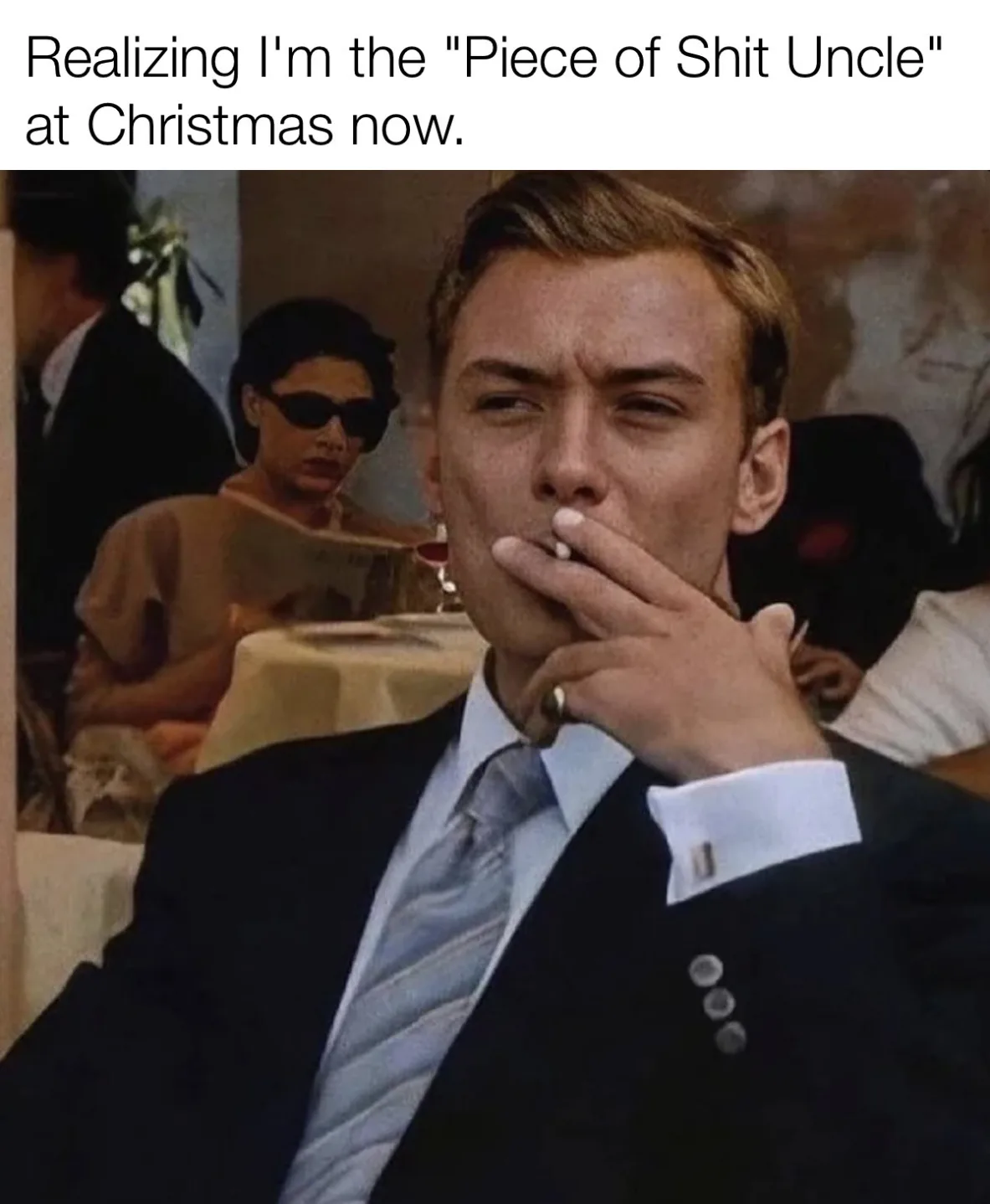 A man in a dark suit and light blue tie holds a cigarette to his lips with a pensive, slightly grimacing expression. Behind…