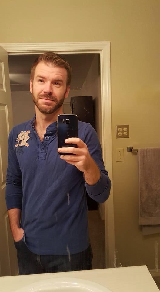 A man with a beard and short hair takes a mirror selfie in a bathroom, holding a black smartphone in his right hand while…