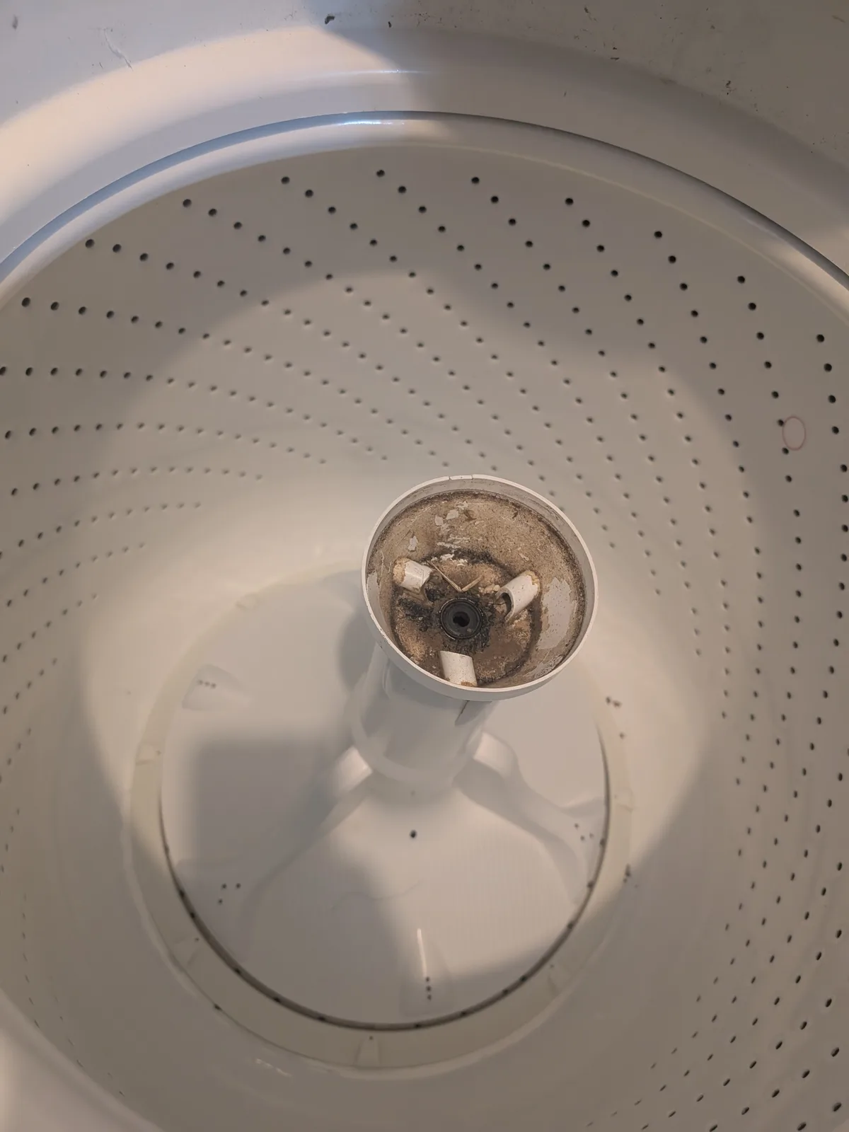 A top-down view of a white washing machine drum showing a heavily clogged central agitator with visible grime, lint, and…