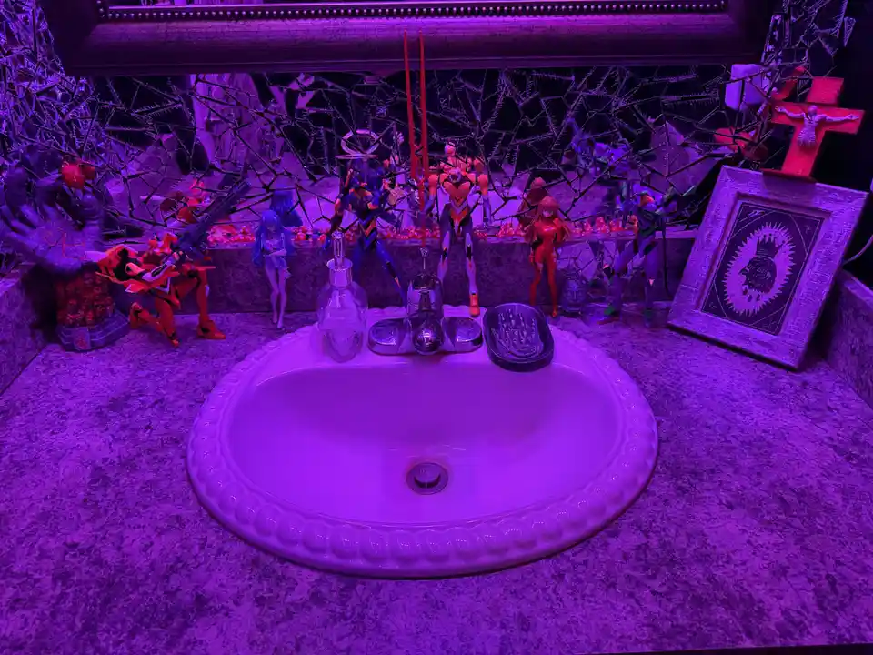 A bathroom sink area bathed in purple light, surrounded by anime figurines, a skull soap dispenser, a framed artwork, and a…