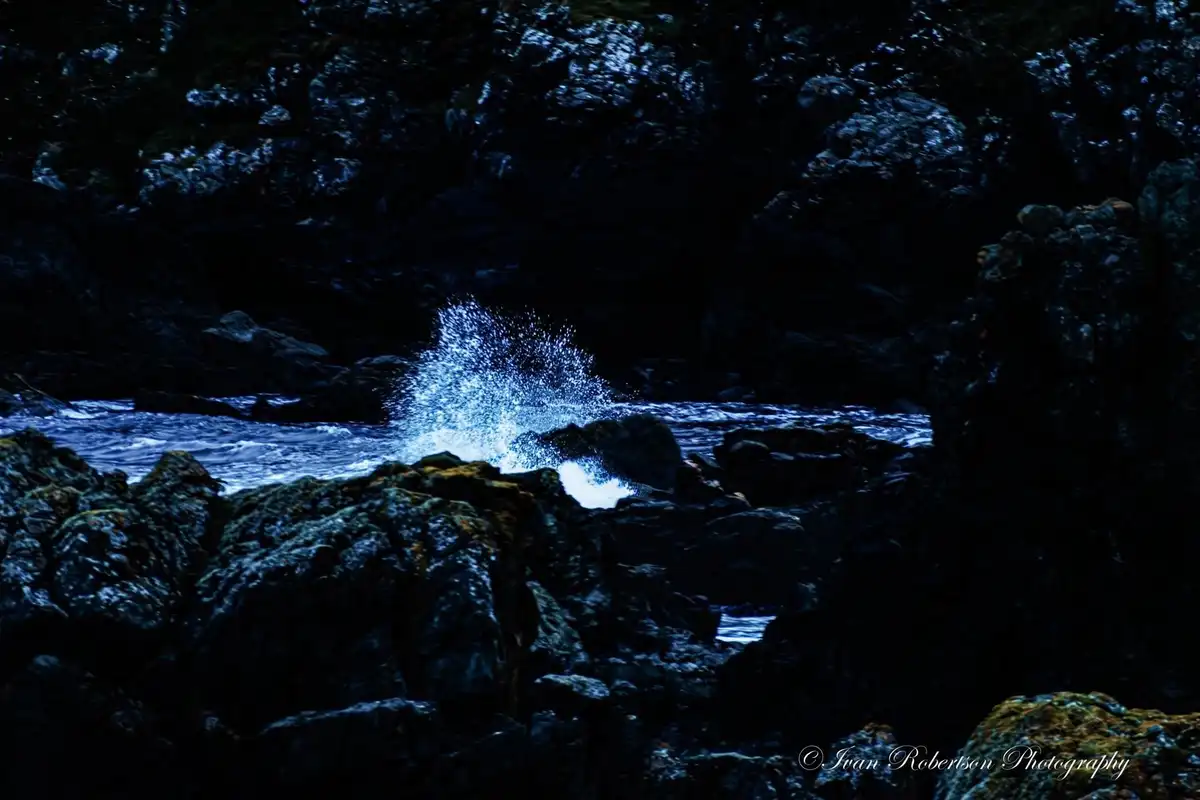 A dark, moody photograph of ocean waves crashing against jagged rocks, creating a bright blue splash of water in the center…