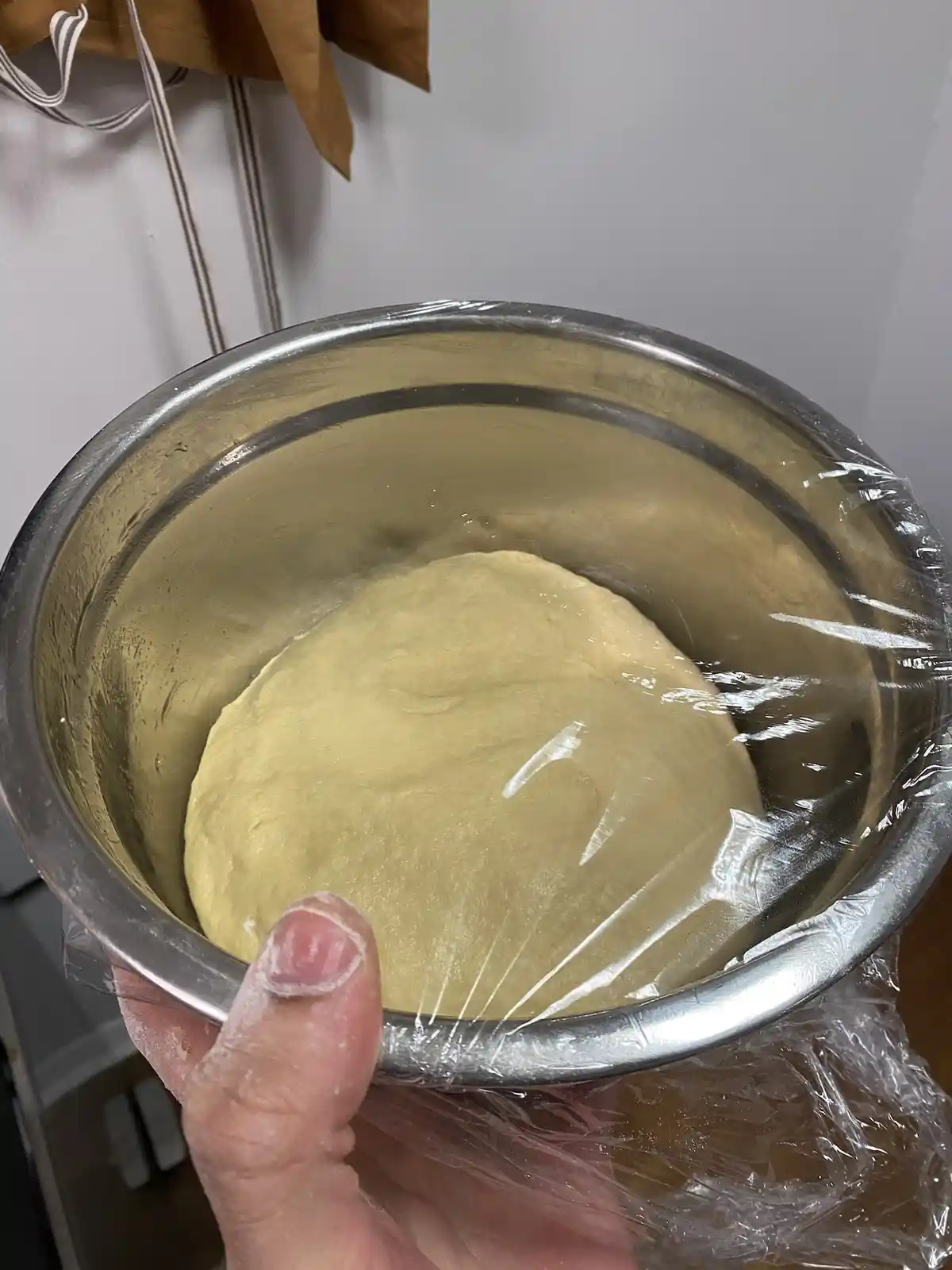 A hand holds a metal mixing bowl containing pale yellow dough covered with clear plastic wrap. The dough appears unrisen and…
