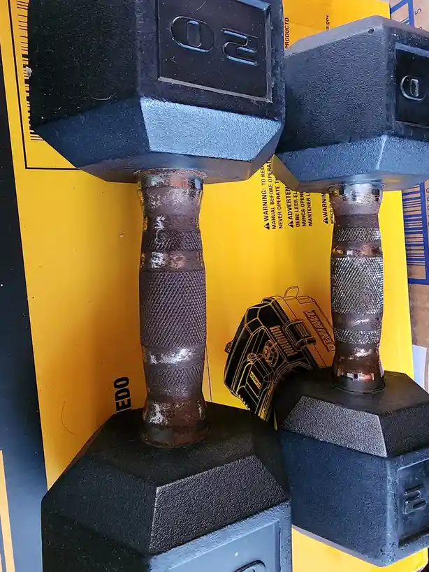 Two 20-pound dumbbells are positioned side-by-side, their handles rusted and textured, resting on a yellow DeWalt box that…