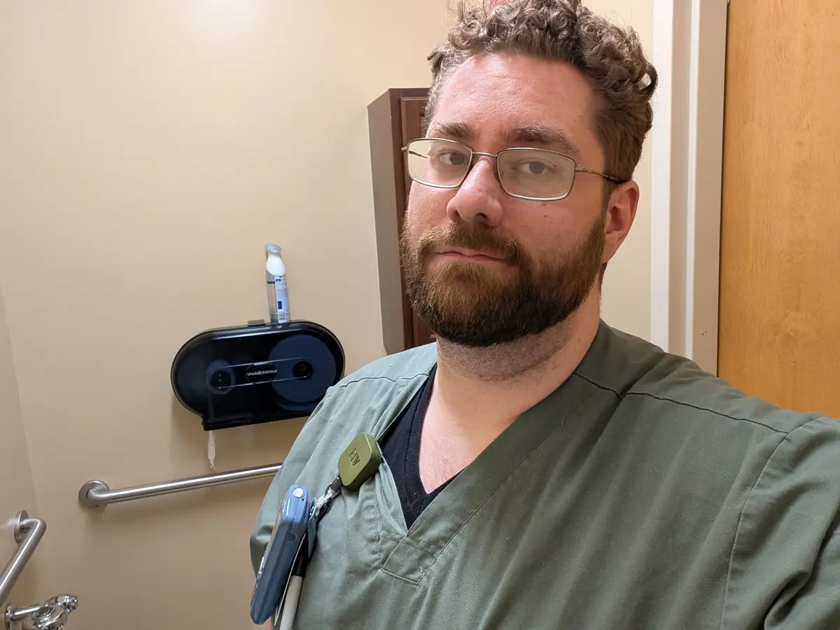 A man with curly hair, glasses, and a beard wearing green scrubs and a name badge stands in a bathroom. He has a serious…