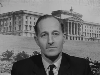 A black-and-white photo shows a man in a suit and tie looking directly at the camera with a serious expression. Behind him…