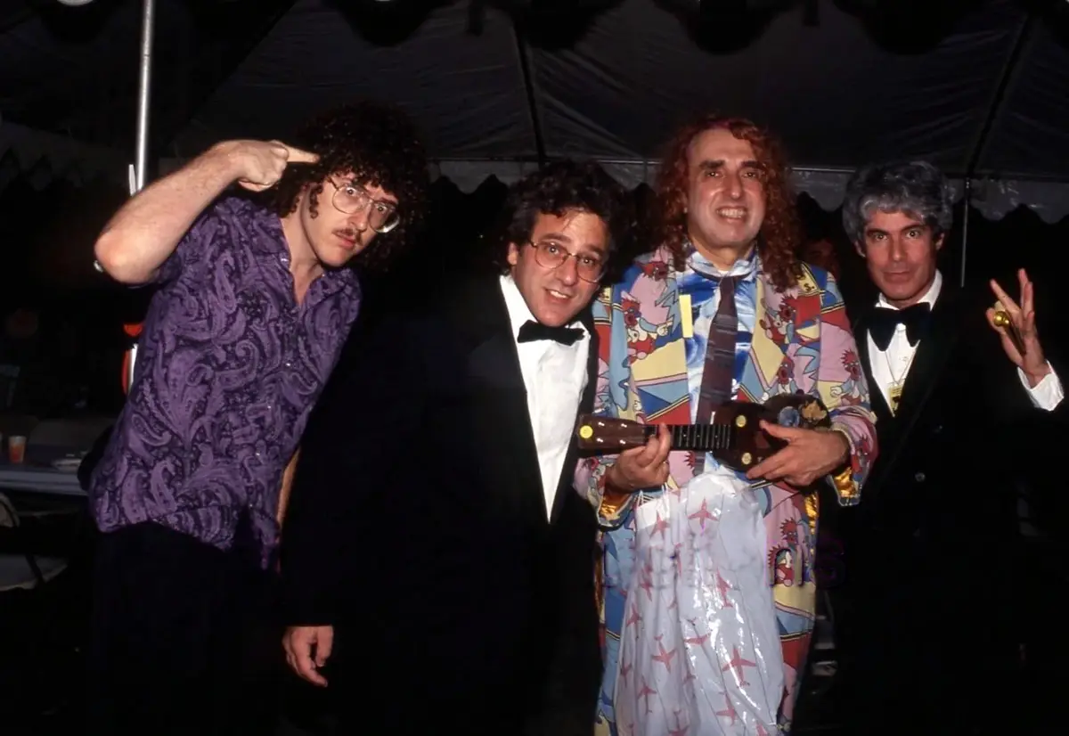 Four men in flamboyant 1980s attire pose together under a tent; one points at his head, another holds a ukulele wrapped in a…