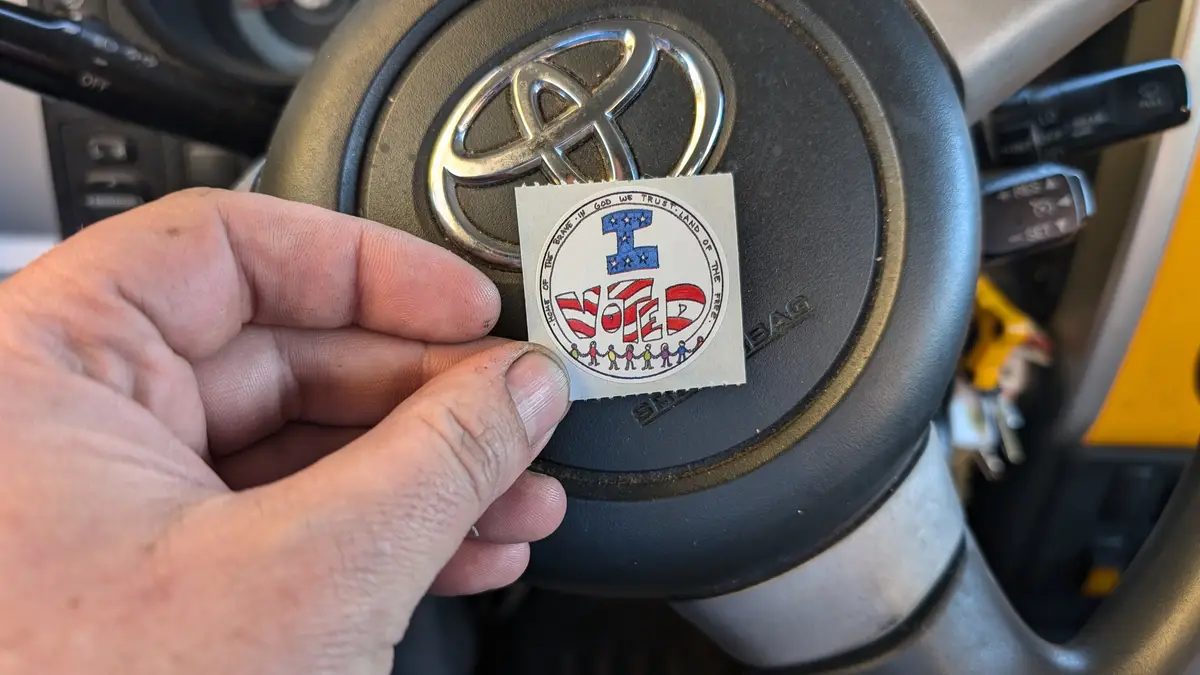 A hand holds an 'I VOTED' sticker in front of a Toyota steering wheel, partially obscuring the logo. The sticker features…