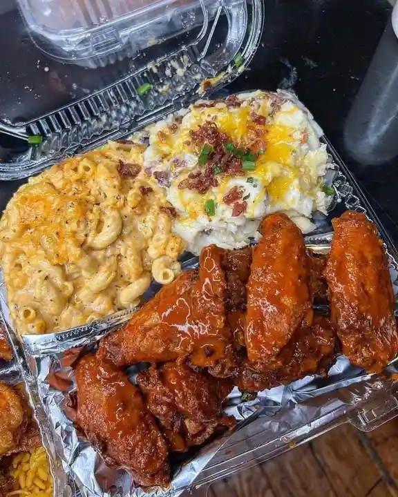 A close-up photo of a takeout meal in a clear plastic container, featuring glossy buffalo wings, creamy macaroni and cheese…