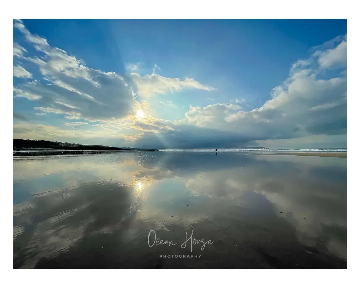 A serene beach scene with a wide, wet sandy shore reflecting a dramatic sky filled with clouds and sunbeams. A lone person…
