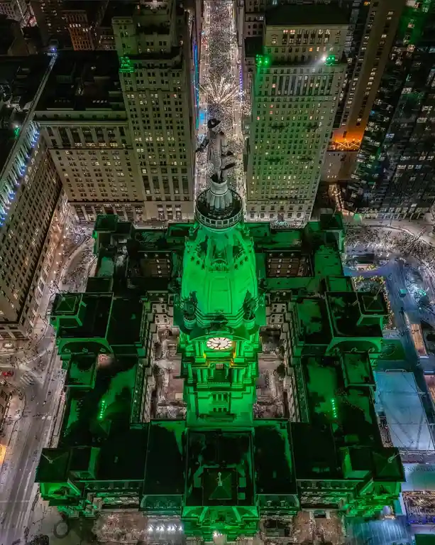 An aerial night photo shows a grand, ornate building illuminated in bright green, with a statue on its roof and city streets…
