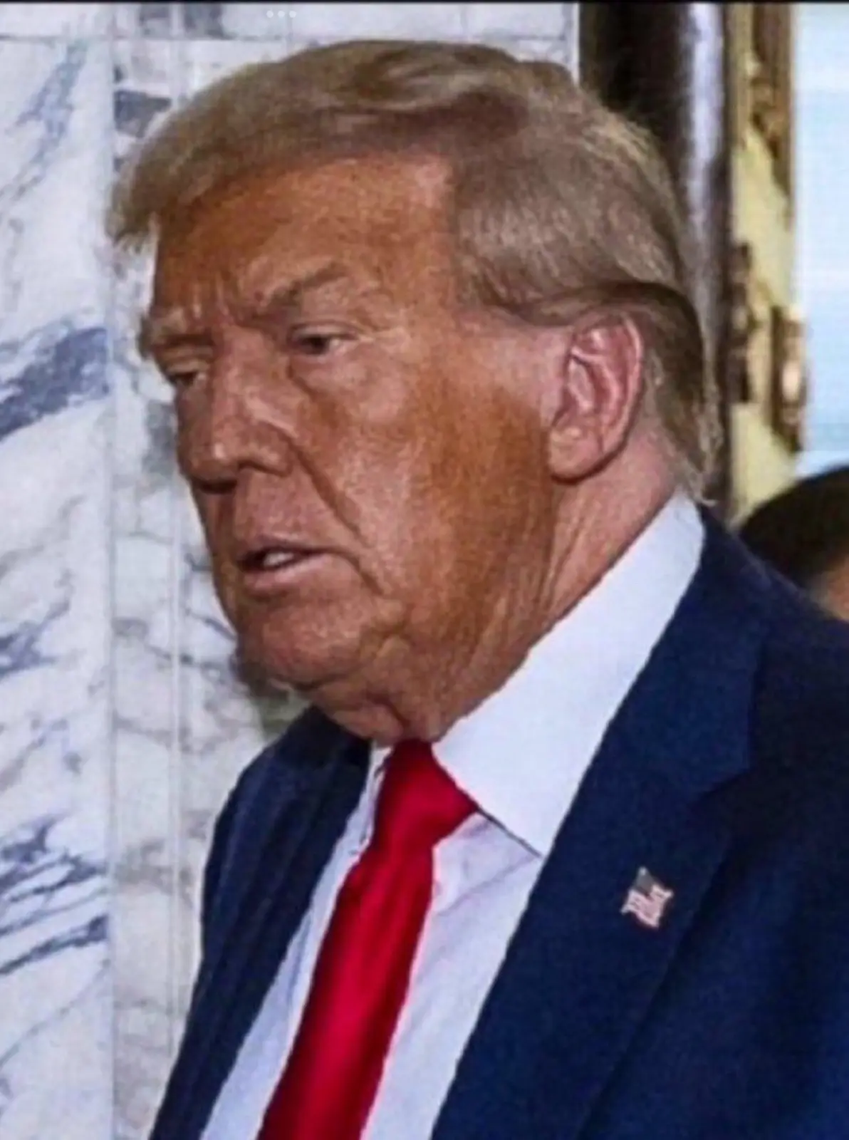 A close-up photo of Donald Trump looking slightly to his left with a serious expression, wearing a dark blue suit, white…
