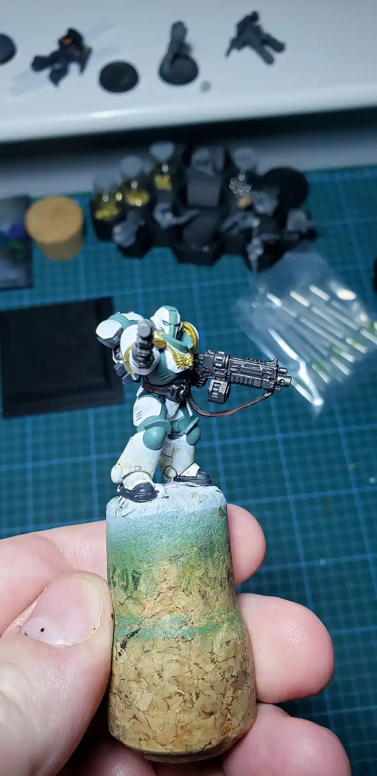 A hand holds a painted miniature of a futuristic soldier in white and teal armor, standing atop a cork stopper that has been…