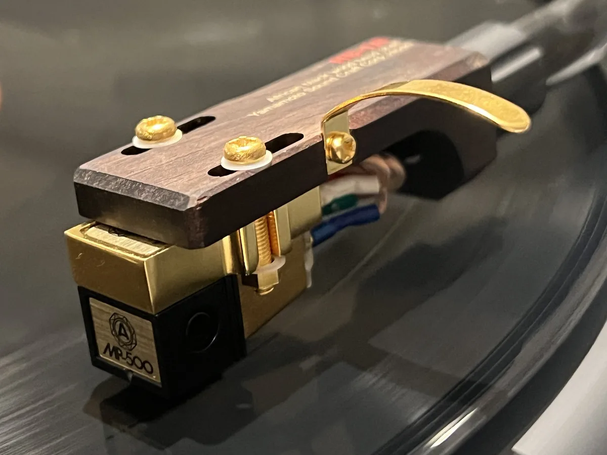 A close-up photo of a high-end, gold-plated turntable cartridge with a wooden top plate and visible wiring, resting on a…