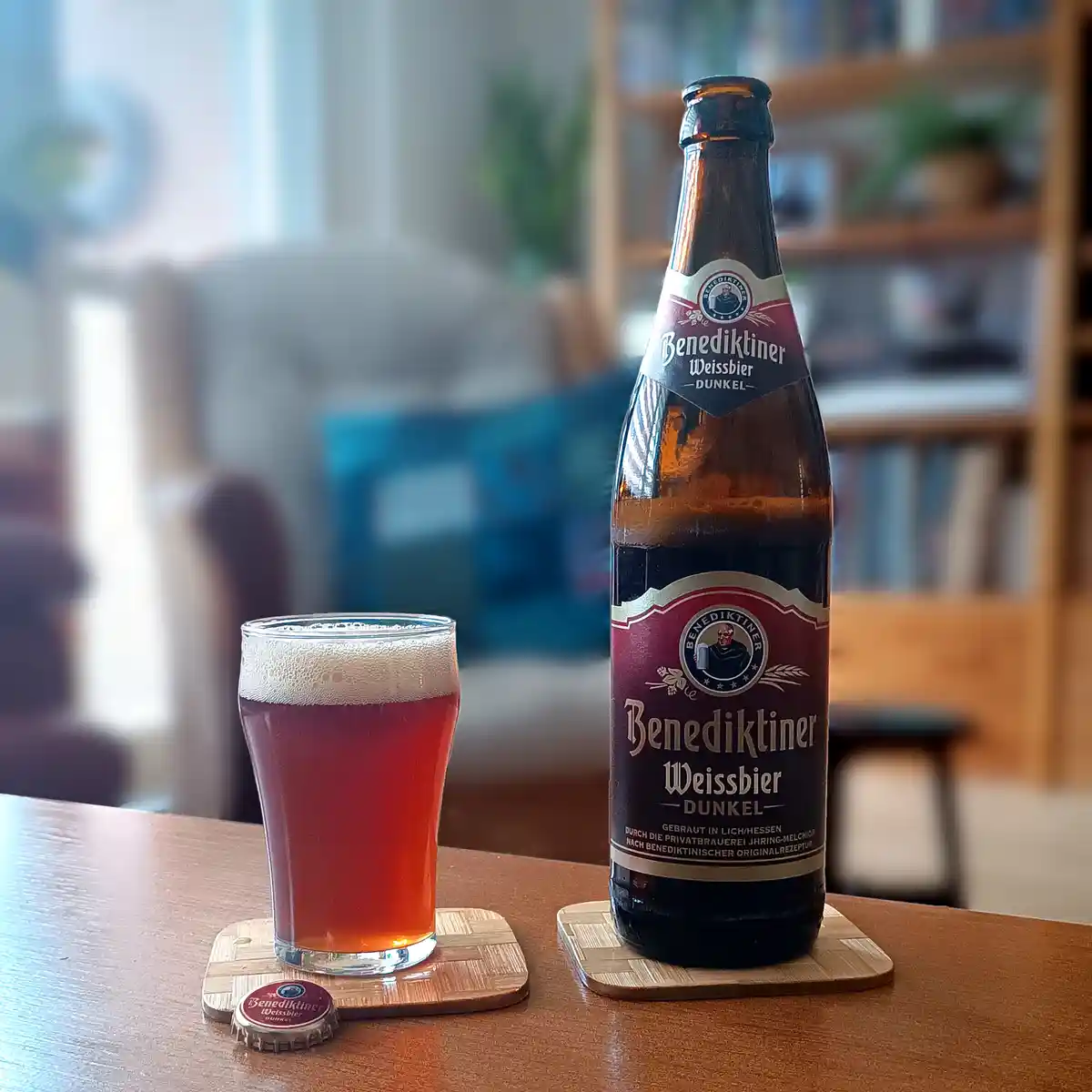A bottle and a glass of Benediktiner Weissbier Dunkel beer sit on wooden coasters on a table, with the bottle's label…