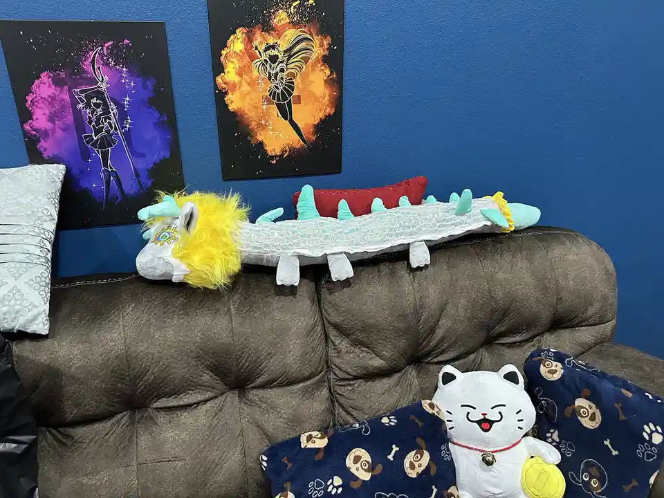 A long, white plush dragon with a yellow mane and turquoise spikes rests horizontally across the back of a brown couch…