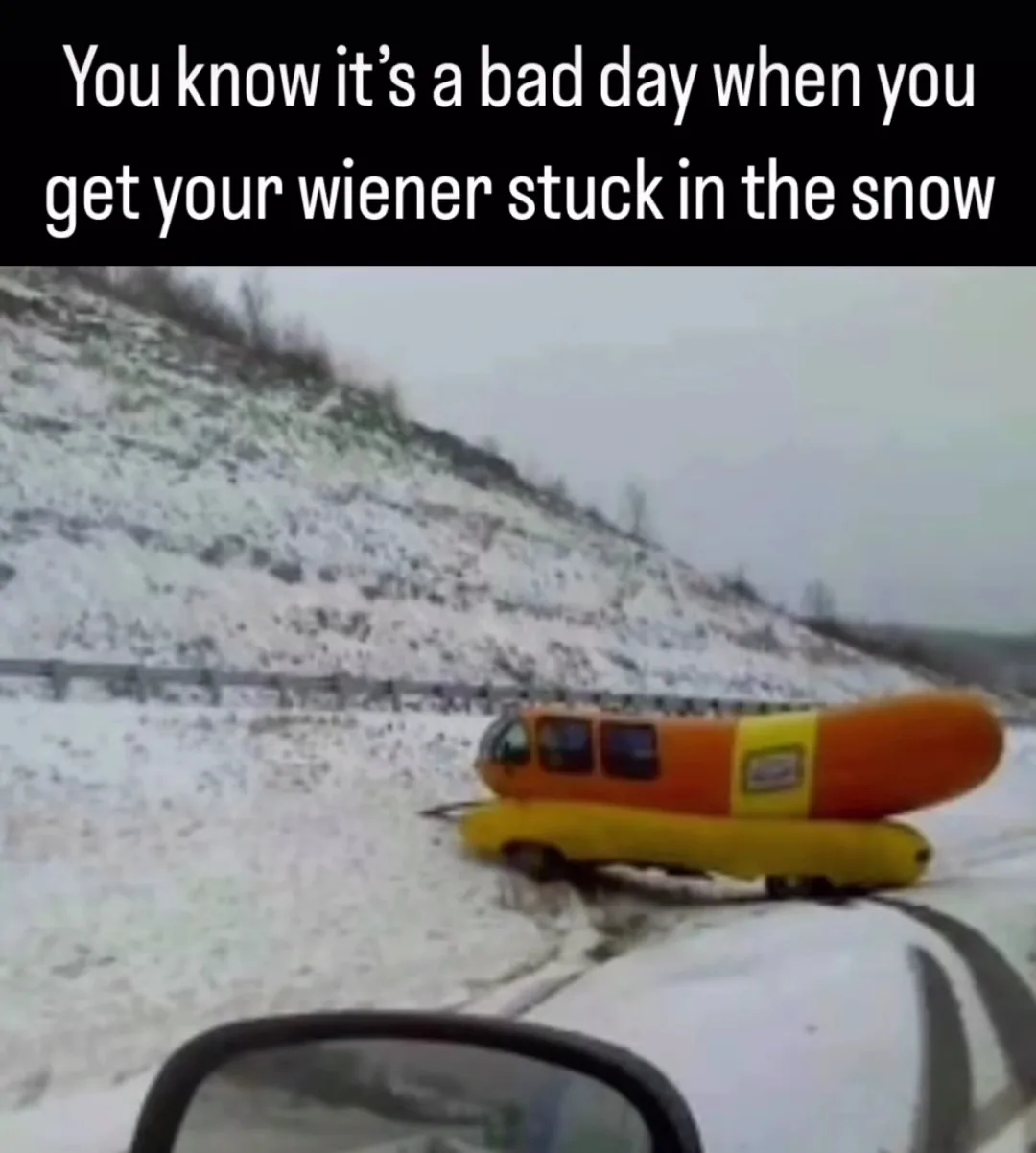 A snowy roadside scene shows a small, orange-and-yellow vehicle shaped like a hot dog (wiener) partially stuck in the snow…