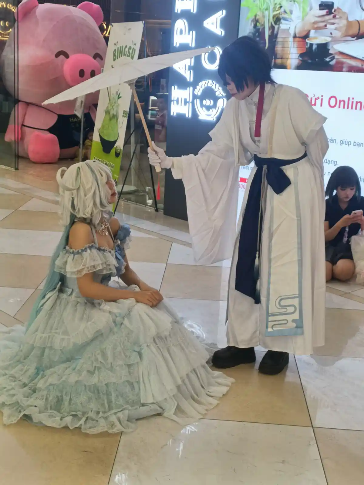 Two cosplayers in a mall: one in a white robe holding an umbrella over a seated person in a frilly blue dress. The standing…