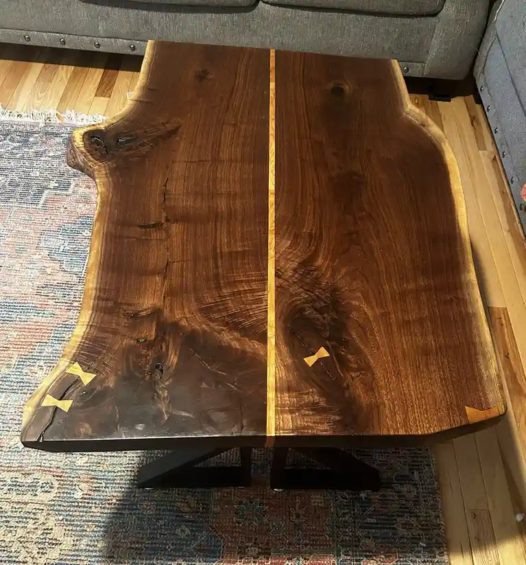 A top-down photo of a rustic live-edge wooden coffee table with a central seam and two small bowtie-shaped inlays, one on…