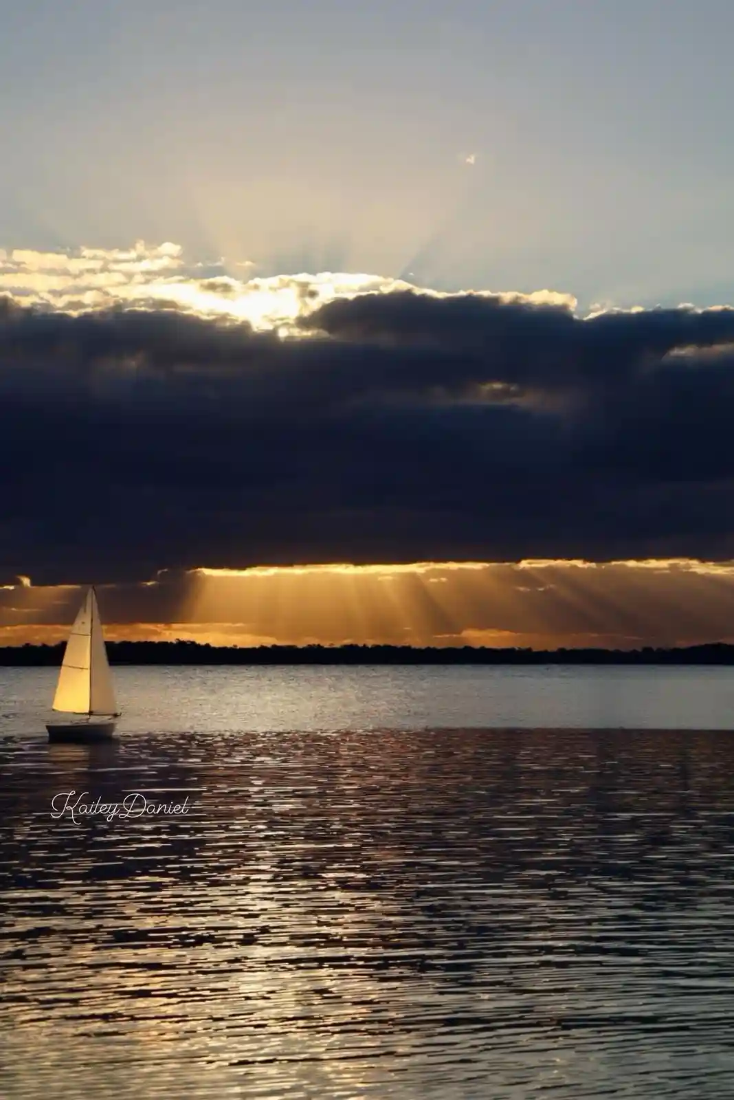 A serene photograph shows a single sailboat on calm water under a dramatic sky where golden sun rays pierce through dark…