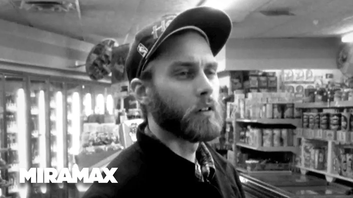 A black-and-white image of a bearded man wearing a baseball cap, standing in what appears to be a convenience store with…