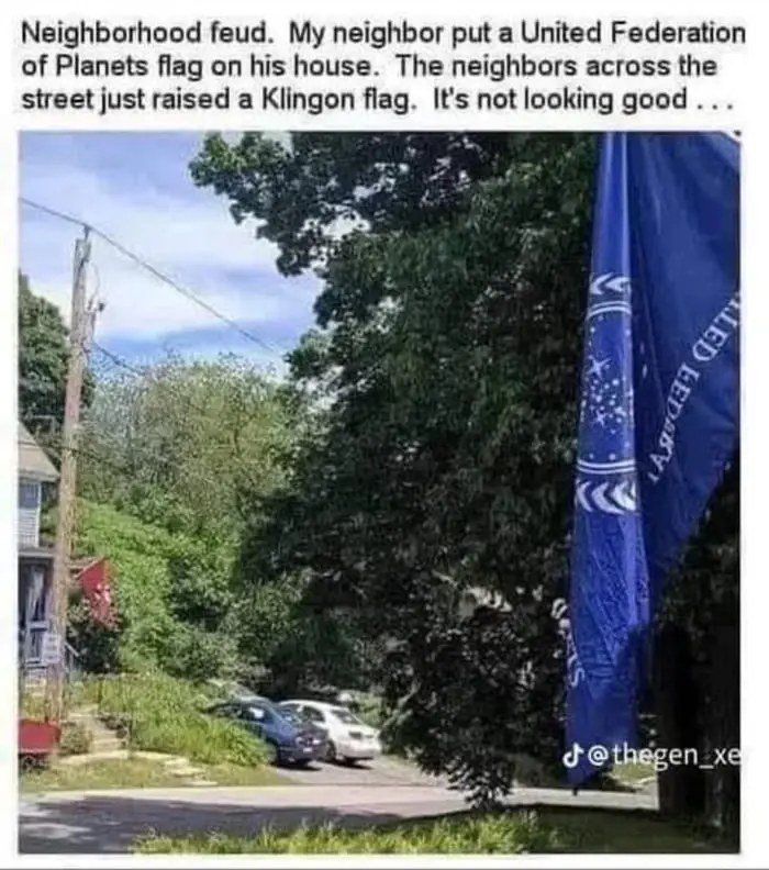 A photo shows two residential houses across a street, each with a flagpole. The house on the right has a large blue flag…