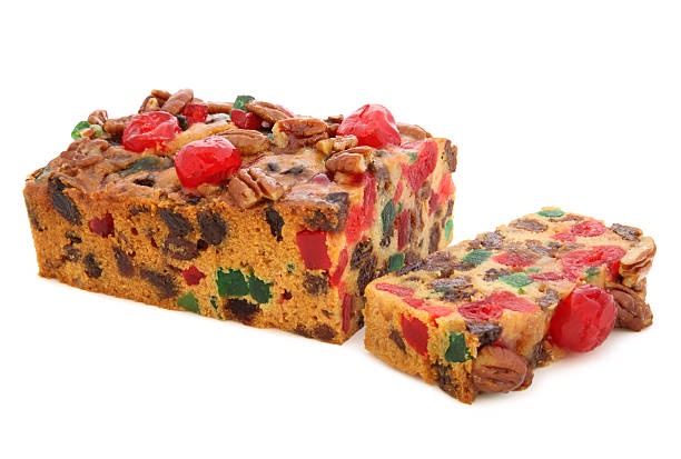 A large rectangular fruitcake with a slice cut from it, both pieces topped with candied cherries, nuts, and colorful candied…