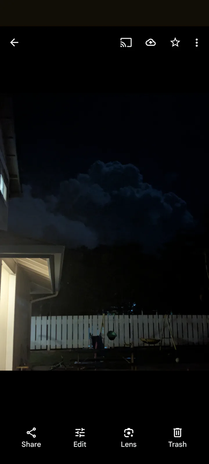 A nighttime photo shows a large, dark cloud looming over a backyard with a white fence and swing set, with the overlaid text…