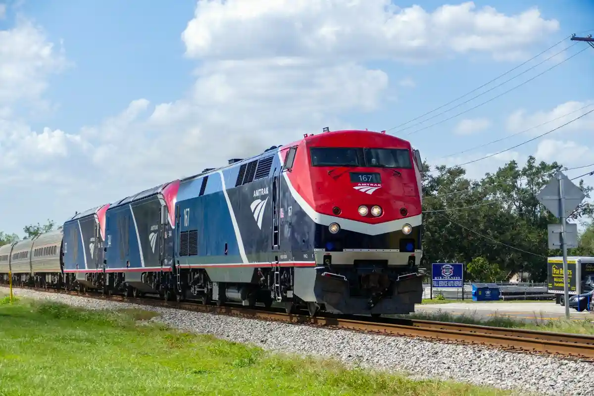 An Amtrak train, locomotive number 167, is shown traveling on tracks under a partly cloudy sky. The train has a red, blue…