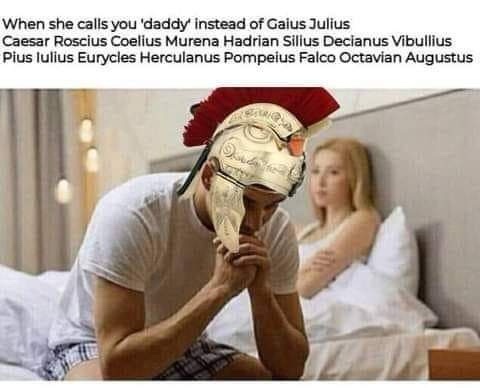 A man wearing a Roman-style helmet sits on a bed with his head in his hands, appearing distressed, while a woman lies in bed…