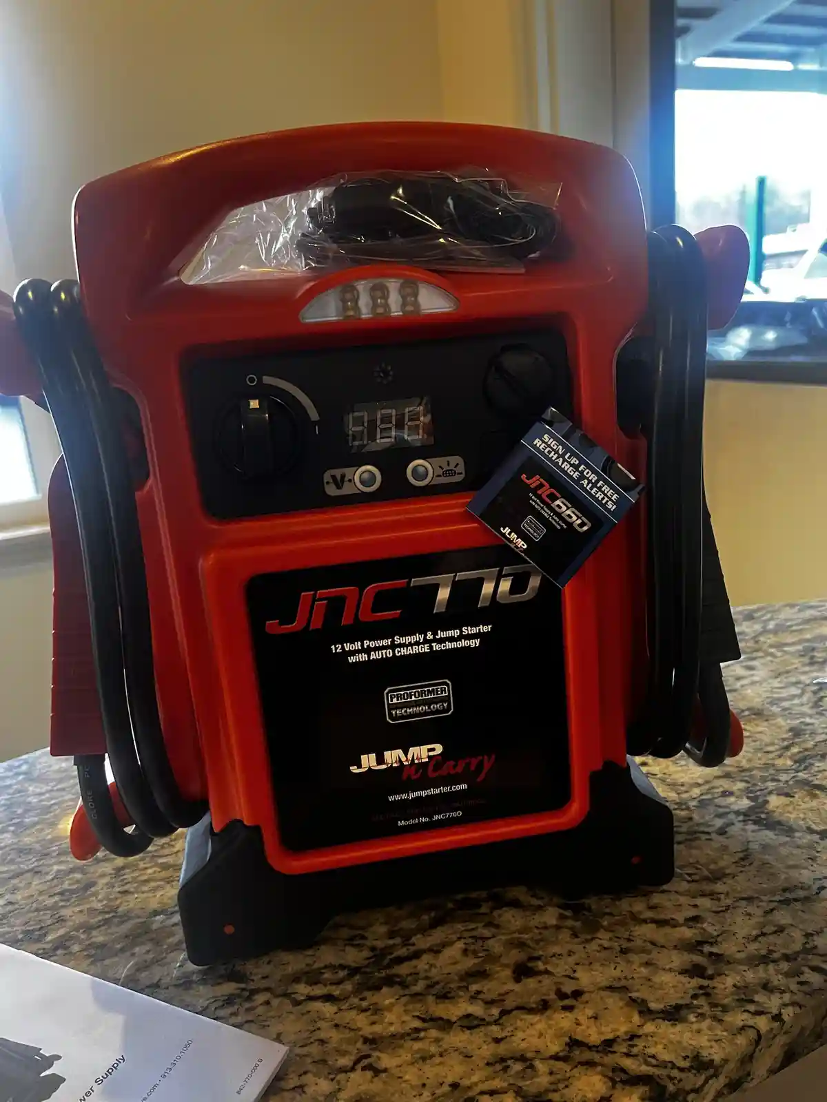 A red JNC770 portable jump starter with black cables sits on a granite countertop, its digital display reads '0.00'. A tag…