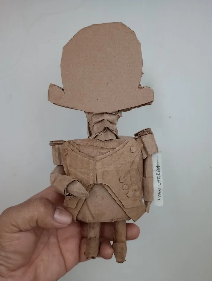 A hand holds a small, crudely crafted figure made entirely from brown paper or cardboard, resembling a stylized human with a…