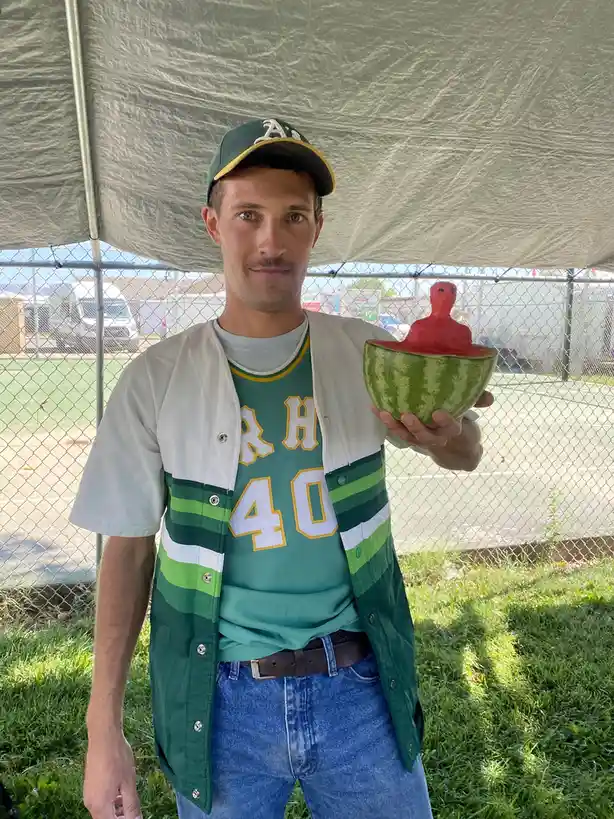 A man wearing a green and white baseball uniform and cap stands under a tent, holding a watermelon that has been carved to…