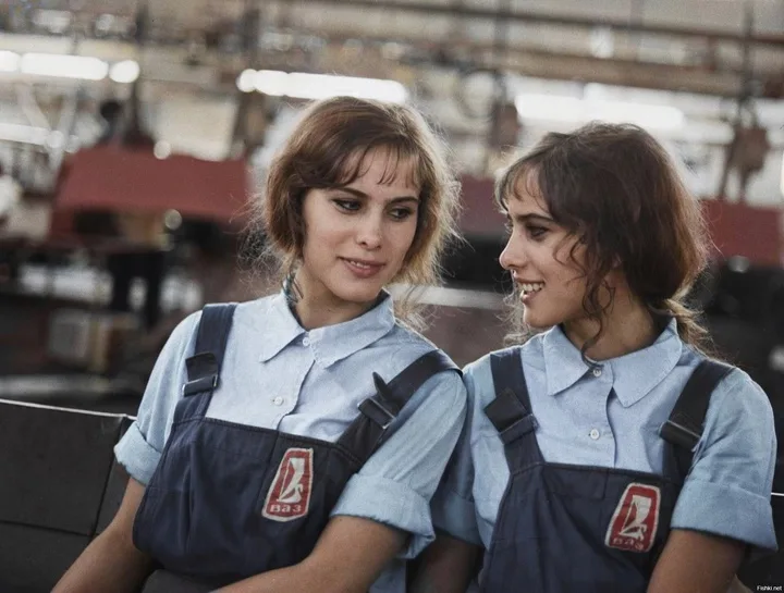 Two women in matching blue work uniforms with red logos sit side by side in an industrial setting, smiling at each other…