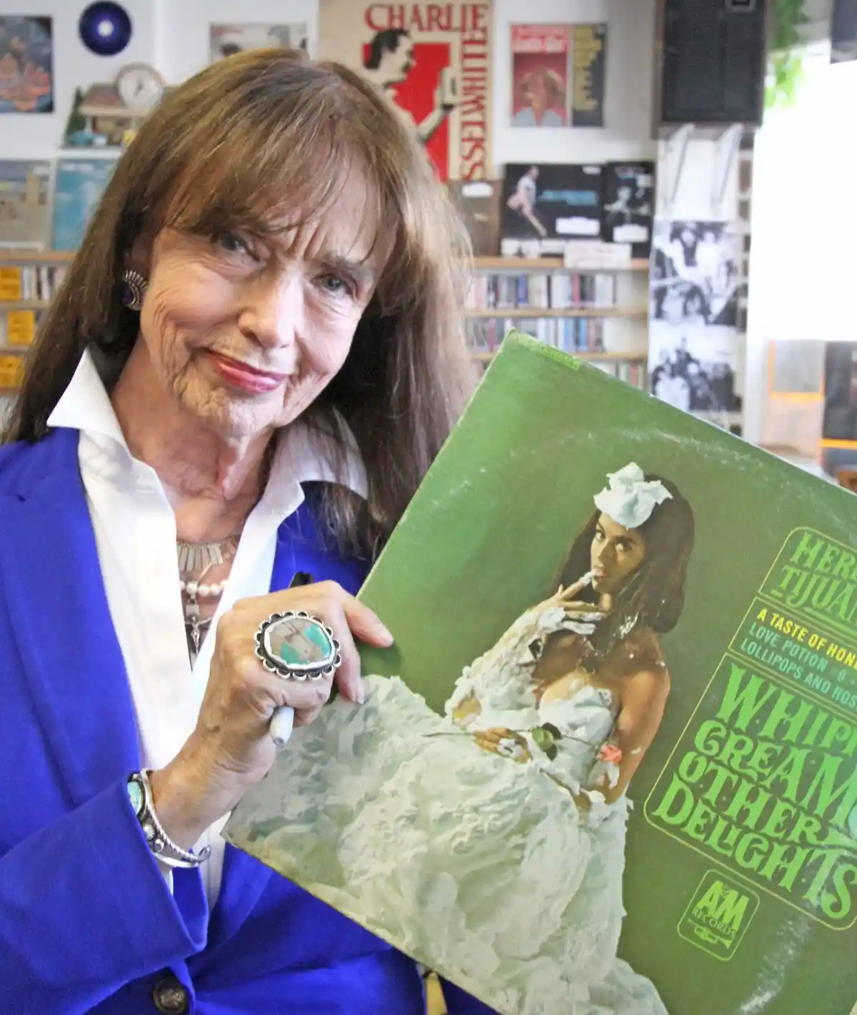 An older woman with brown hair and a blue blazer holds a vintage green album cover featuring a woman in a white dress…