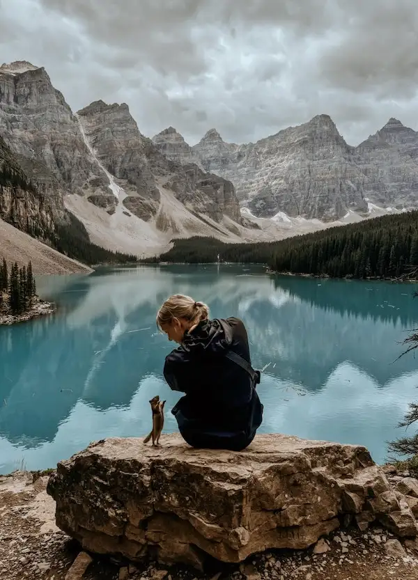 A person in a dark jacket sits on a large rock overlooking a serene, turquoise lake surrounded by towering, snow-dusted…