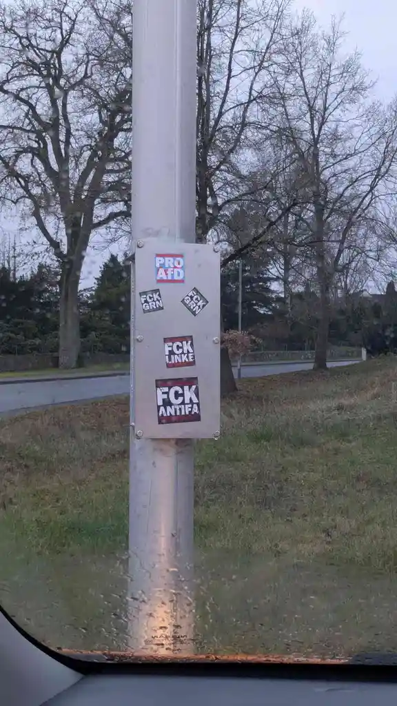 A metal utility box on a pole is covered in political stickers, including 'PRO AFD', 'FCK GRN' (twice), 'FCK LINKE', and…
