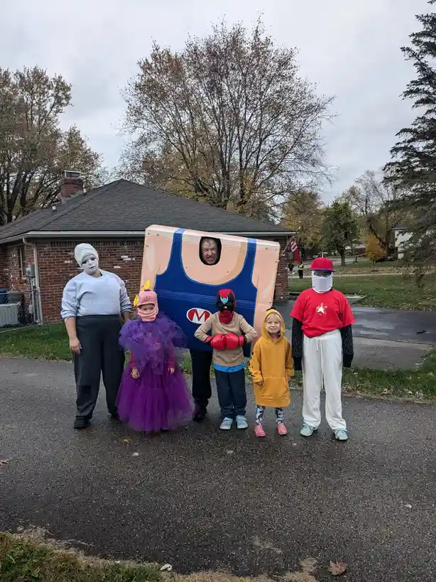 A group of five people, including children, pose for a photo outdoors in Halloween costumes. One person is inside a large…