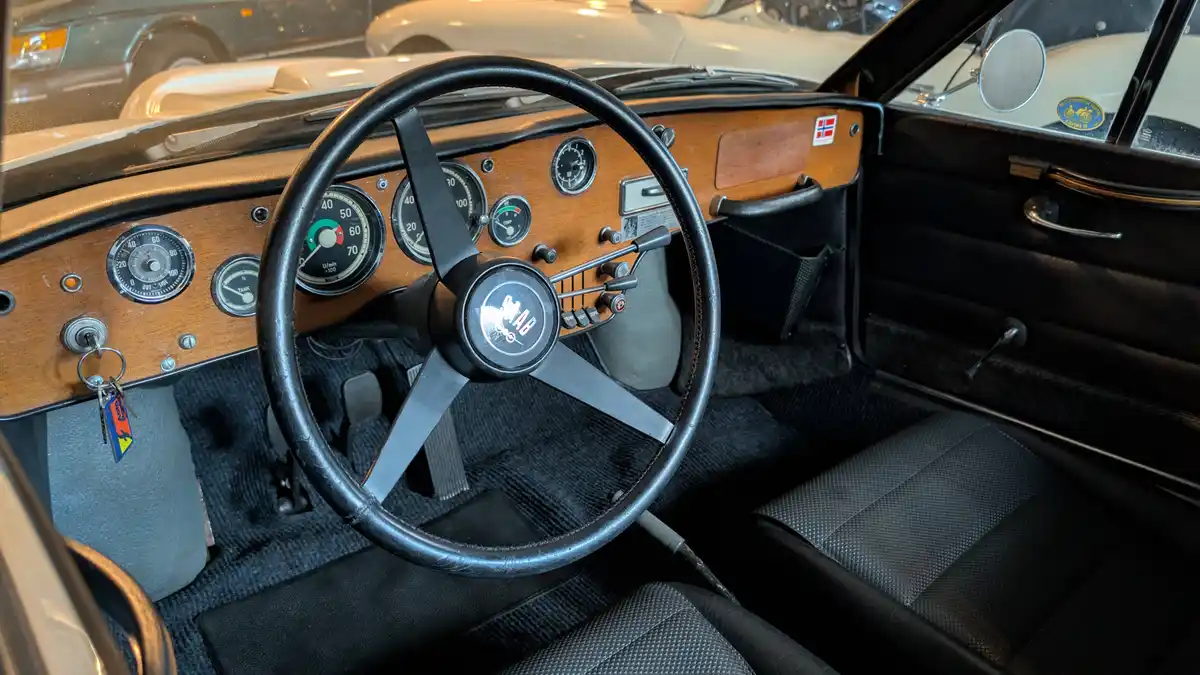 The image shows the interior of a vintage Saab car, featuring a black leather steering wheel with the Saab logo, a wooden…