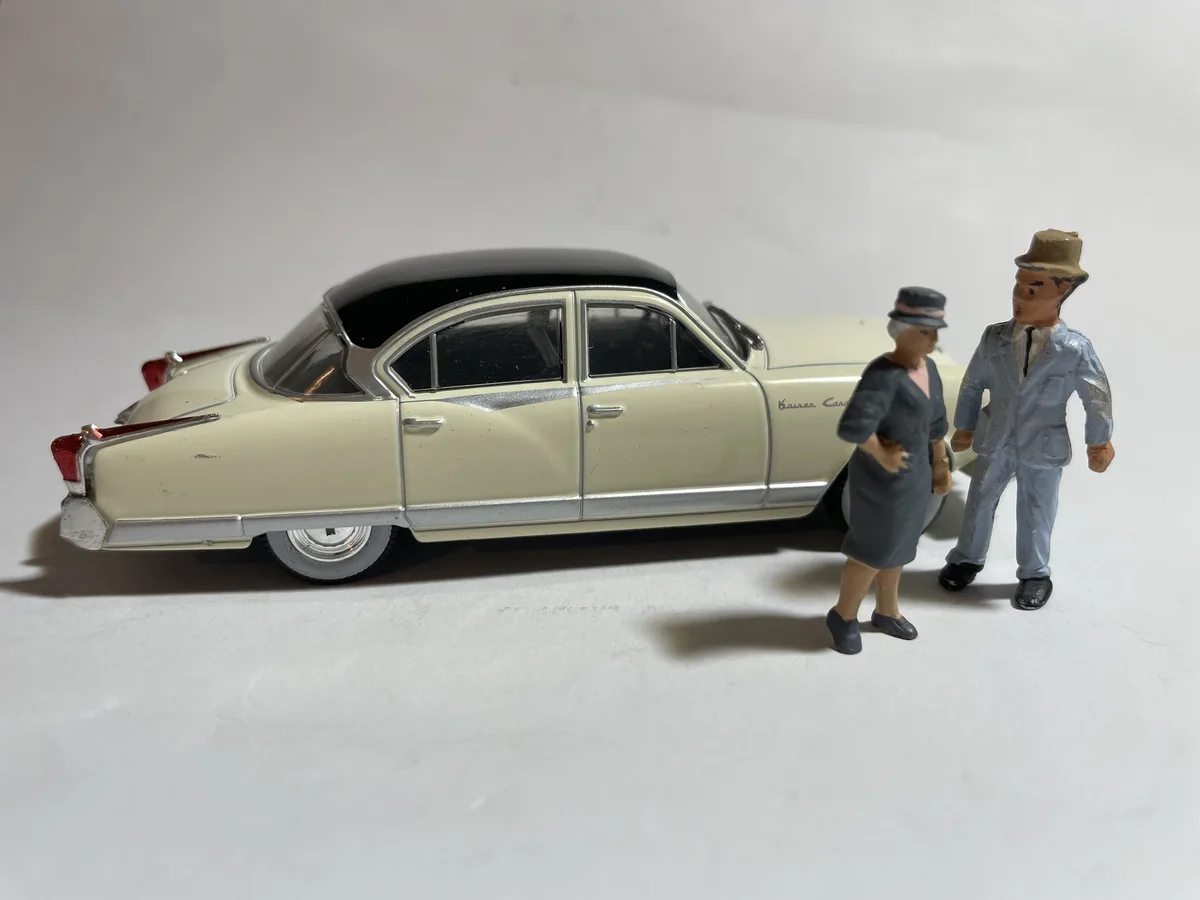 A vintage cream-and-black toy car with 'Kaiser Car' lettering is shown with two small toy figures, a man in a light blue…