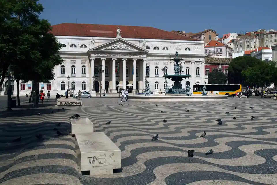 A sunny photo of a grand plaza with a classical building, a central fountain, and a distinctive wavy cobblestone pattern…