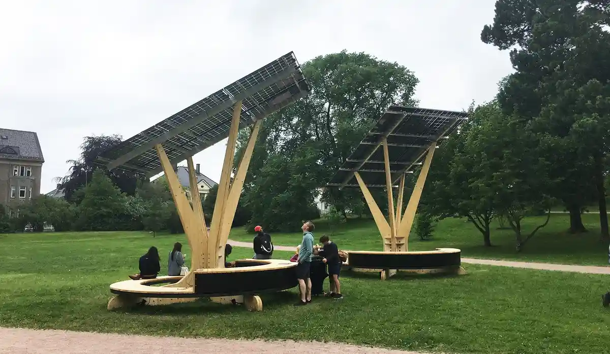 Two large, modern wooden park benches with tilted solar panels above them are shown in a grassy park. Several people are…