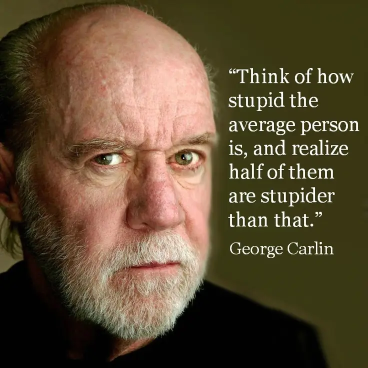 A close-up portrait of George Carlin, a balding older man with a white beard and intense, skeptical expression, looking…