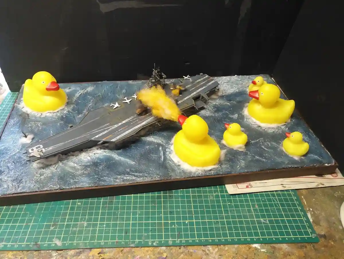 A model of an aircraft carrier is shown on a blue textured surface resembling water, surrounded by several yellow rubber…