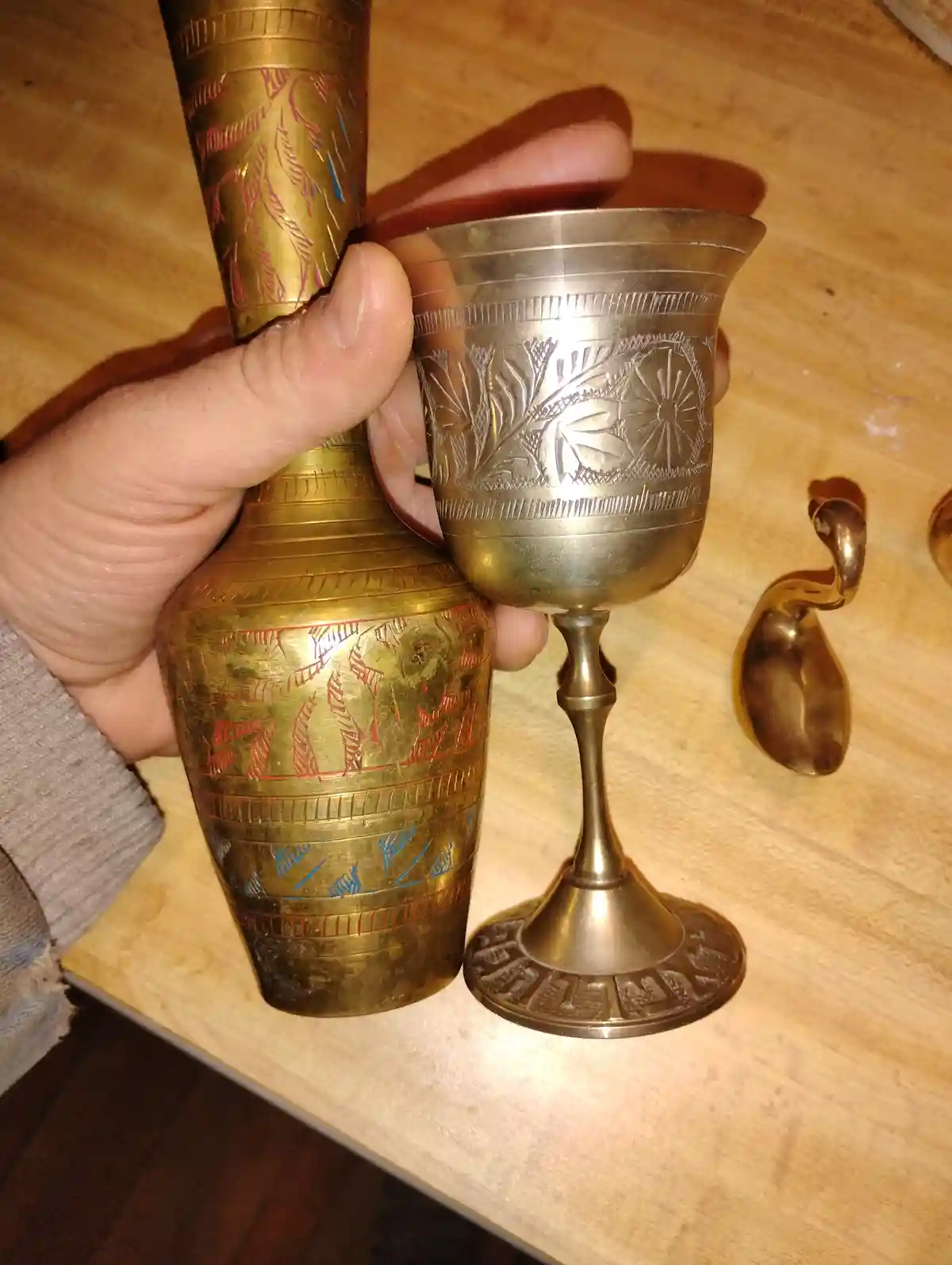 A hand holds a tall, ornate brass vase with red and blue inlaid patterns next to a smaller, engraved brass goblet on a…
